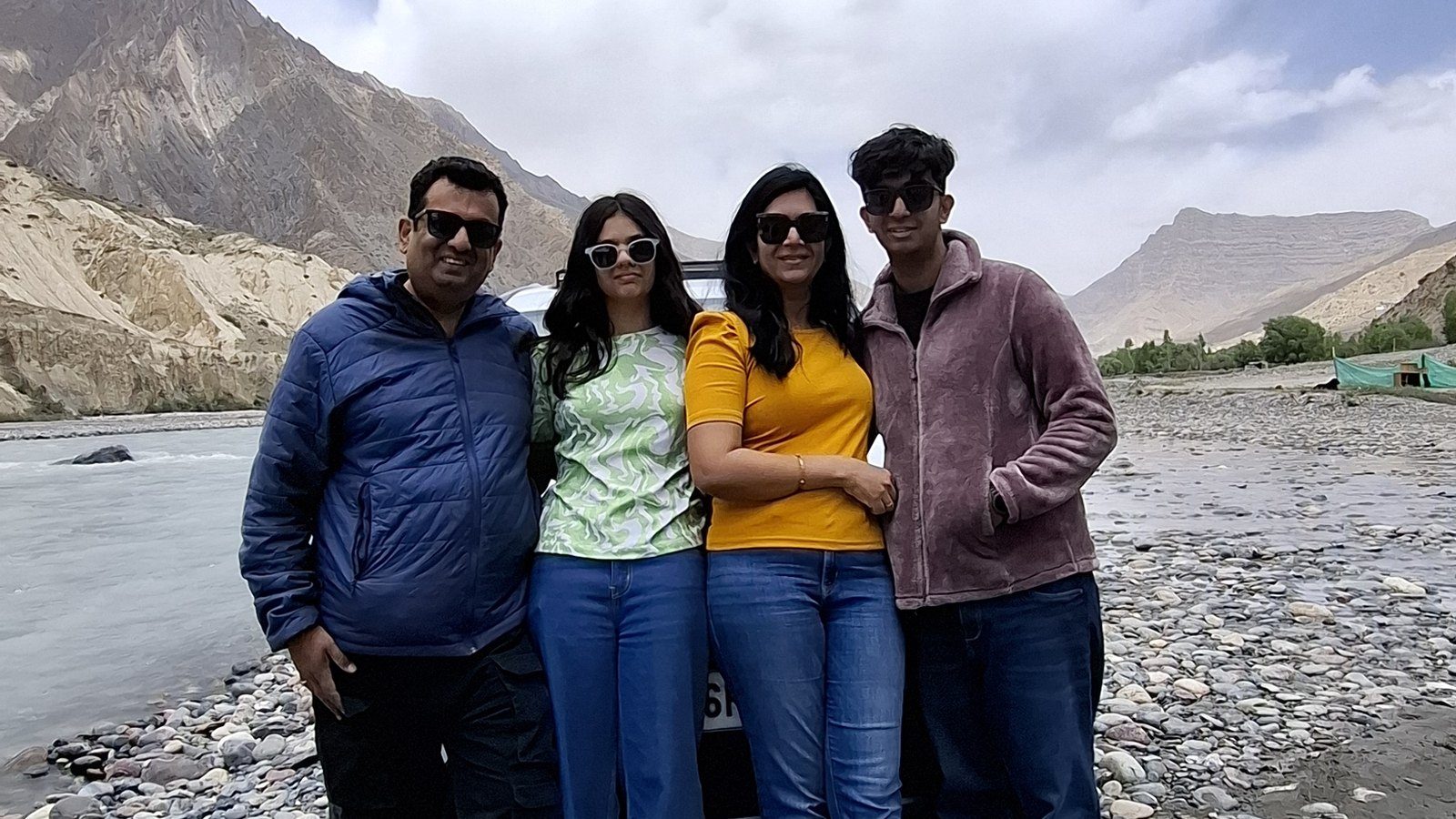 Surabhi Kuthiala's family of four standing together on a stony riverbank high in the mountains, a pale river and bare ridges behind them under a wide cloudy sky.