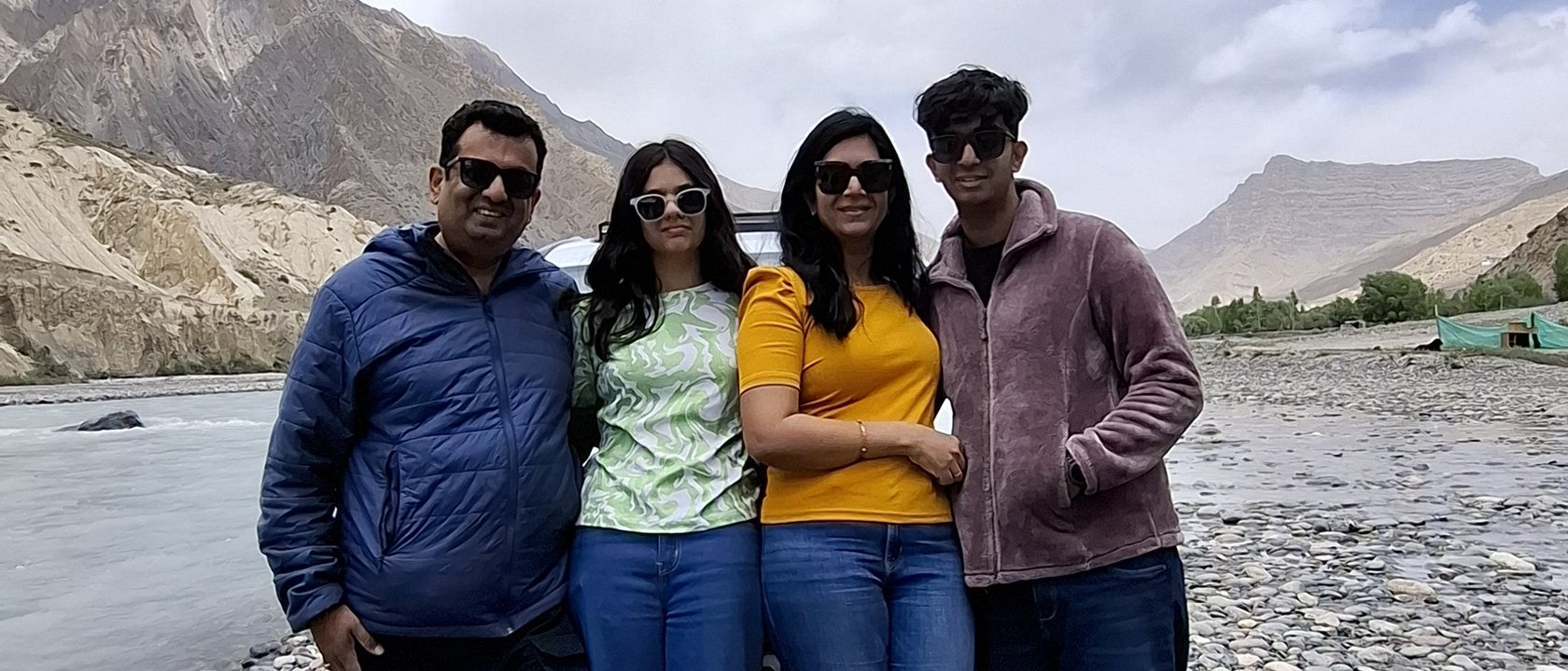 The four members of the family behind CalmNCalculated standing together on a stony riverbank, bare mountain ridges and a pale river behind them.